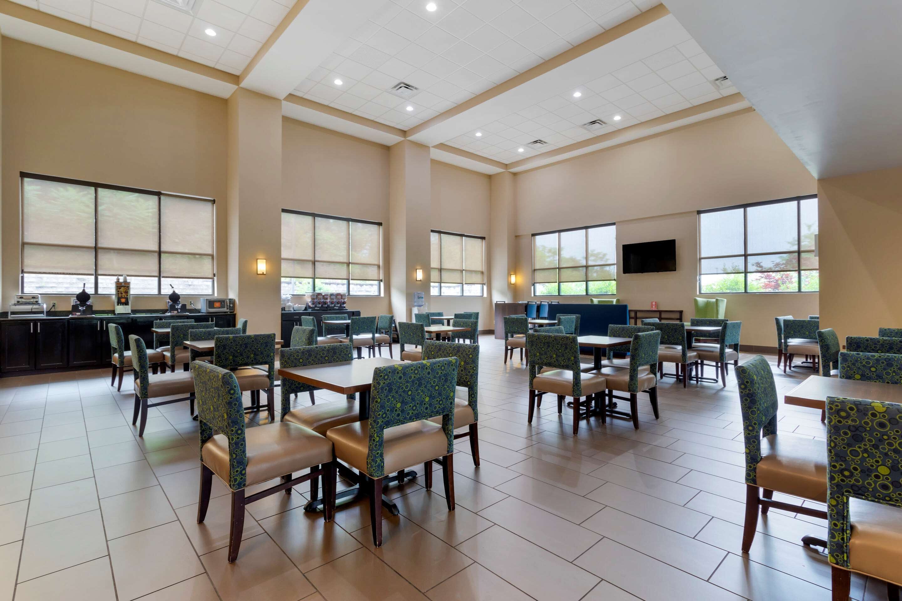 A spacious dining area with multiple tables and chairs, large windows, and a kitchen area in the background.