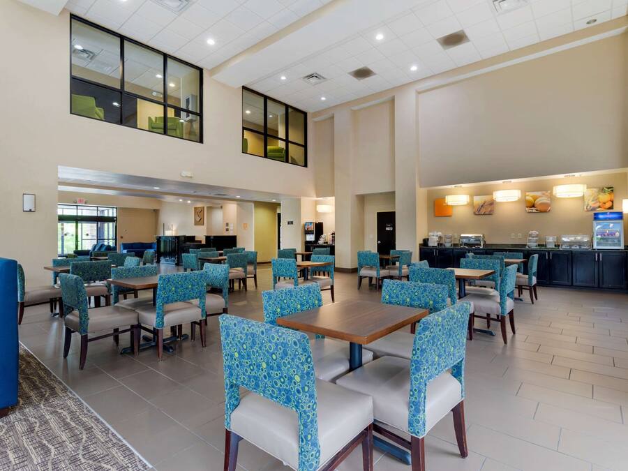 A spacious hotel lobby with multiple dining tables and chairs, a kitchen area, and a view of the outside through large windows.