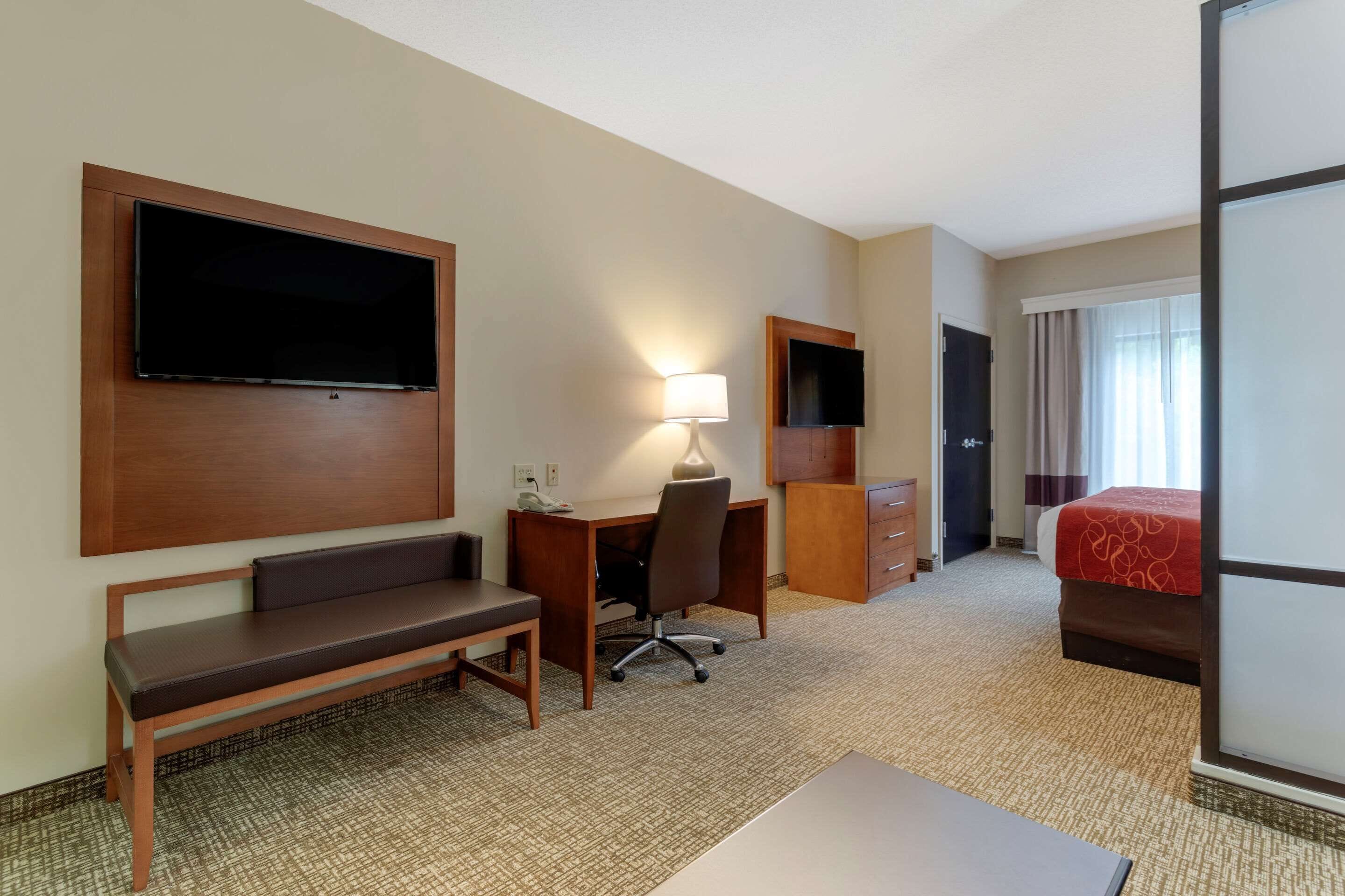 A hotel room with a bed, a desk with a chair, a bench, a TV mounted on the wall, and a lamp.