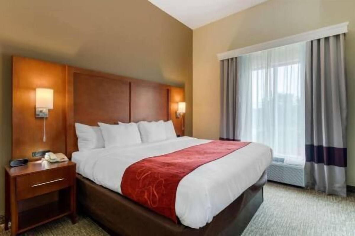 Ein Hotelzimmer mit einem großen Bett, einem Holzkopfstück, Nachttischlampen, einem Beistelltisch und einem Fenster mit Vorhängen.
