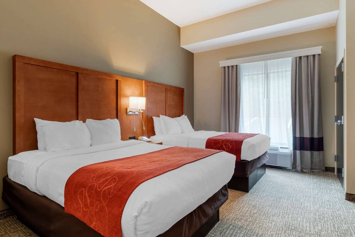 Ein Hotelzimmer mit zwei Betten, einem hölzernen Kopfteil, einem großen Fenster mit Vorhängen und einem Teppichboden.
