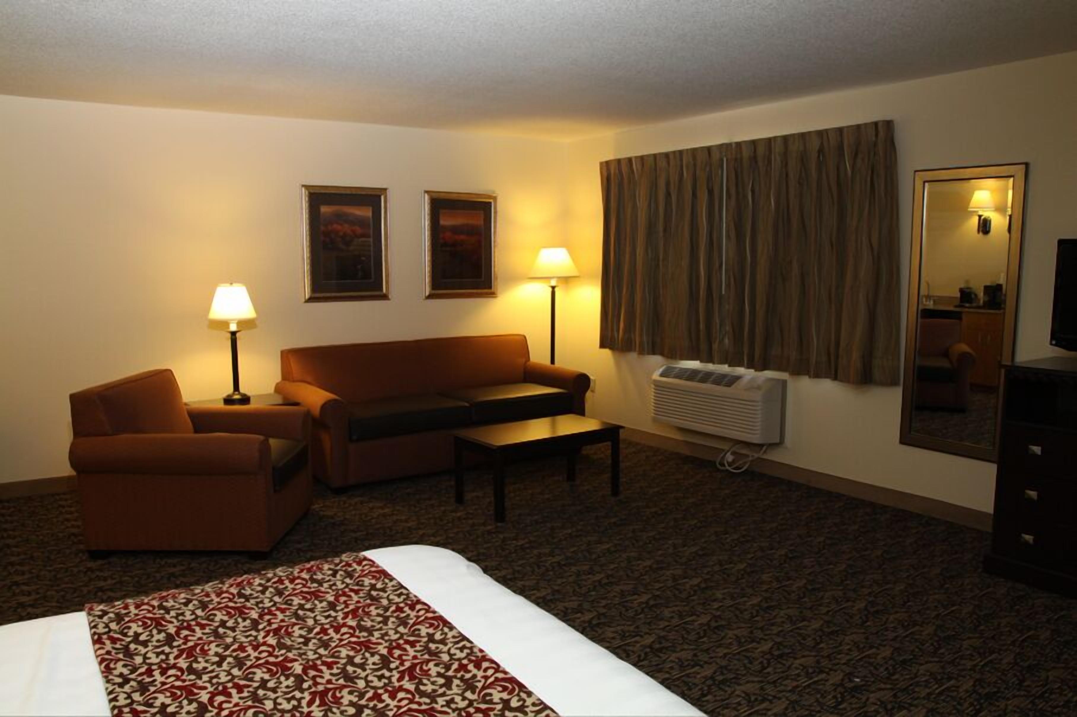 A hotel room with a bed, a sofa, a chair, a table, two lamps, a mirror, and an air conditioning unit.