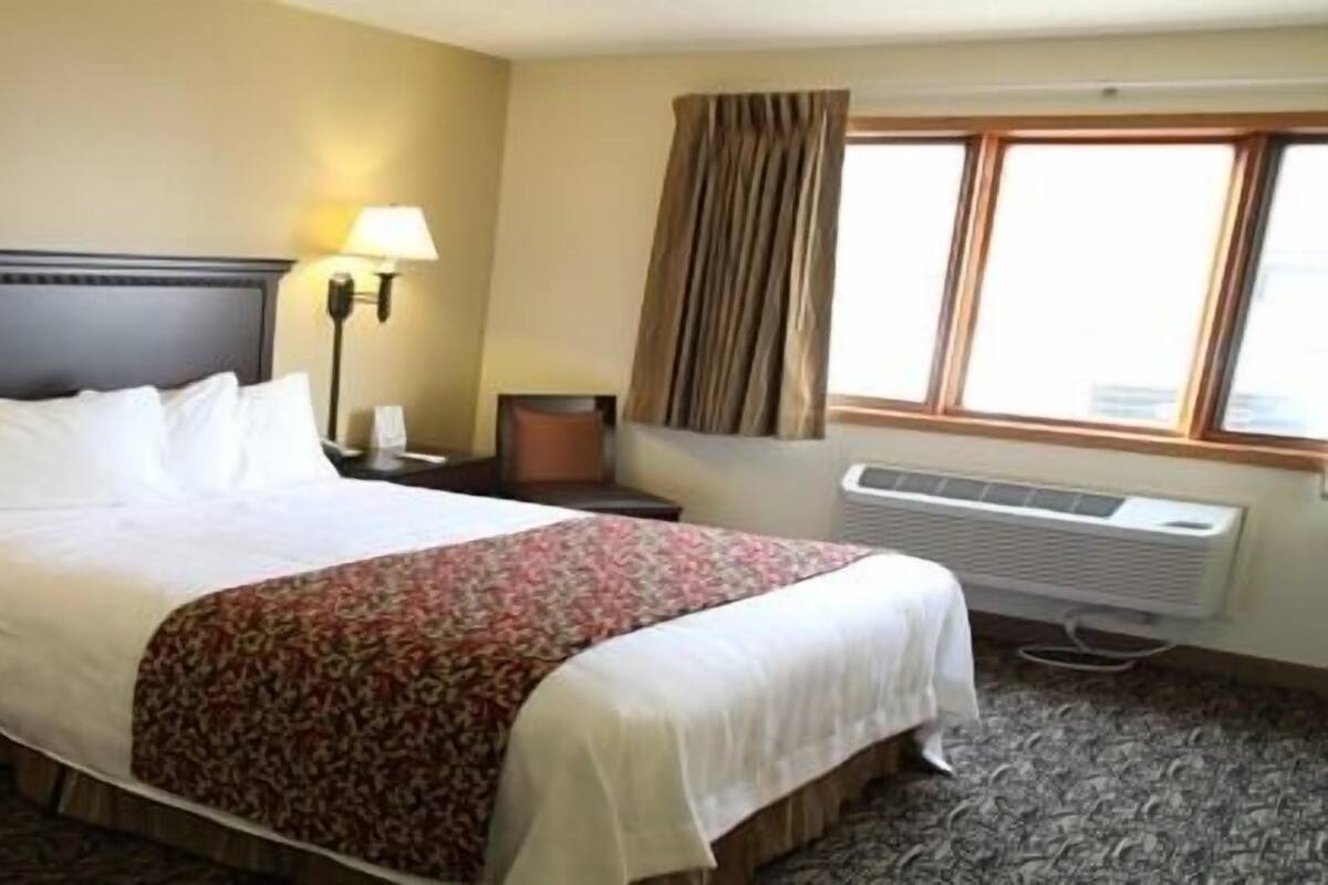 A hotel room with a large bed, two bedside tables, a nightstand, a lamp, a window with curtains, and a wall-mounted air conditioner.