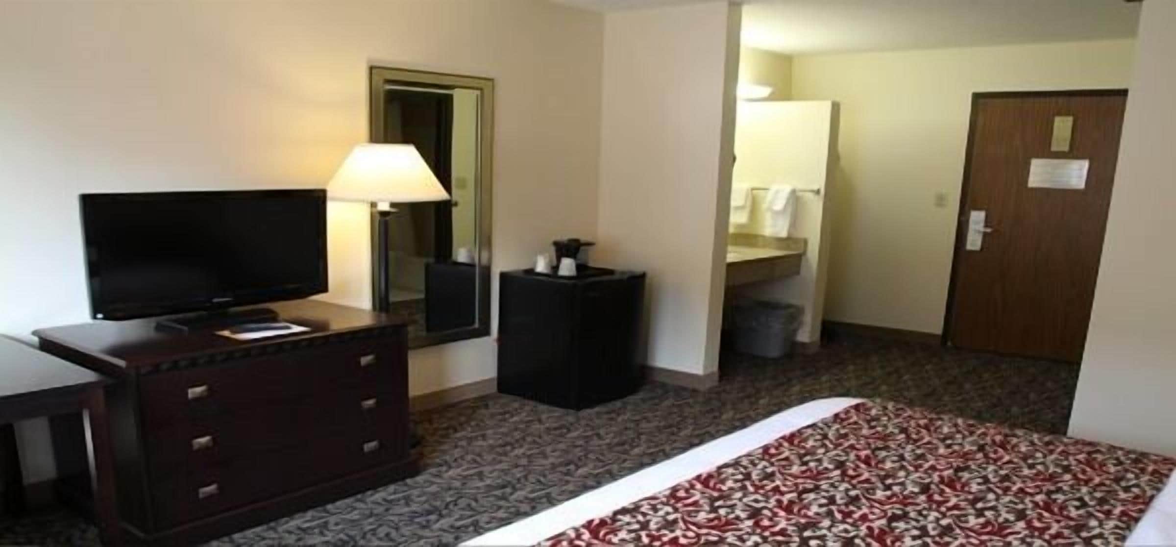Habitación de hotel con una cama, un televisor sobre un tocador, una lámpara, un espejo y una puerta que da a un baño.
