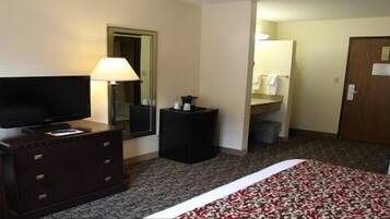 Habitación de hotel con una cama, un televisor sobre un tocador, una lámpara, un espejo y una puerta que da a un baño.