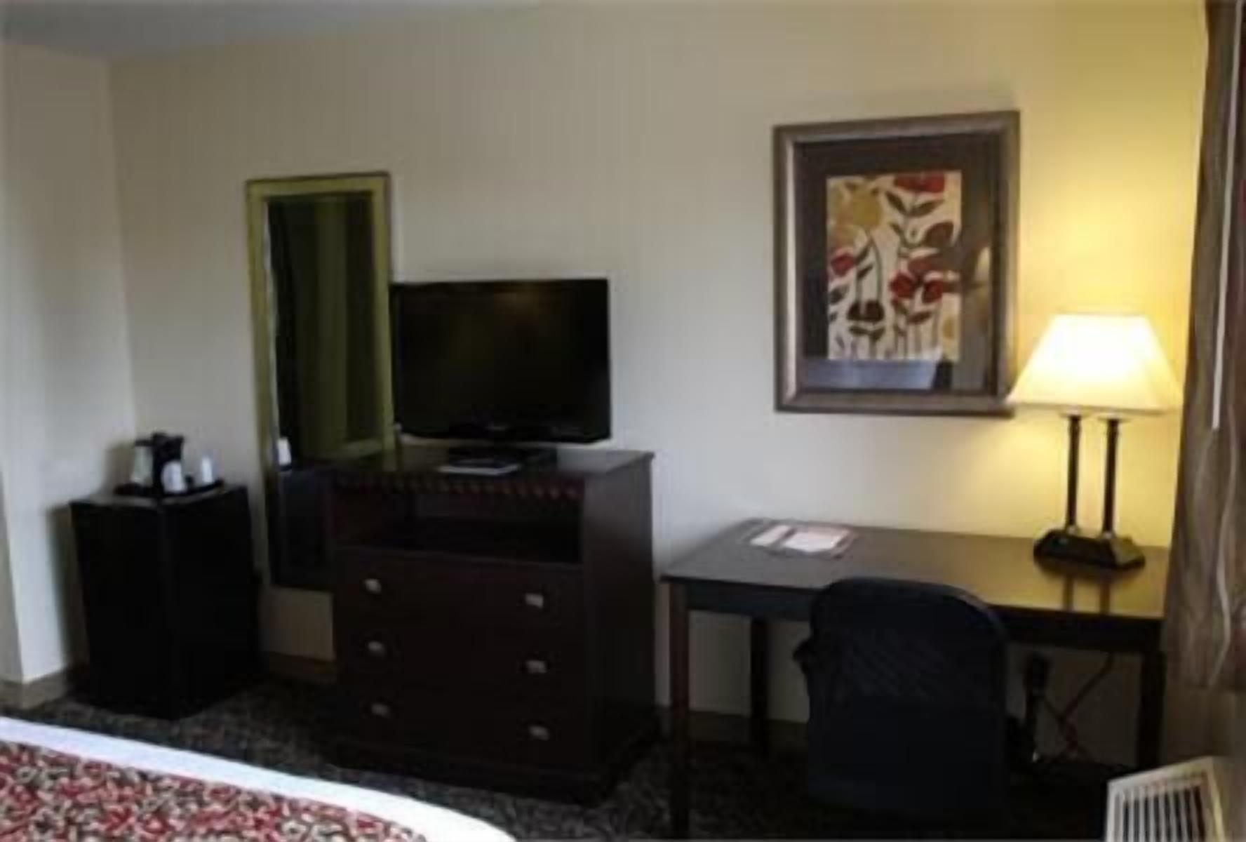 A hotel room with a bed, a television on a dresser, a desk with a chair, a lamp, and a framed picture on the wall.