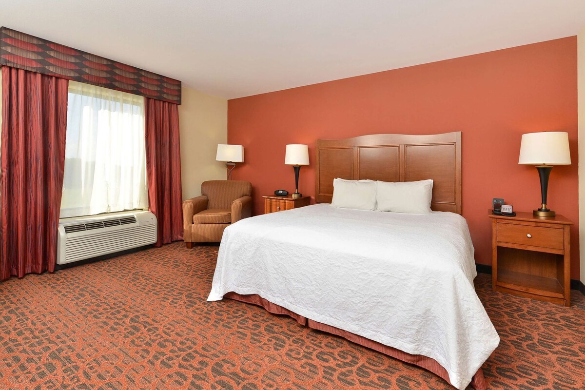 A hotel room with a large bed, two bedside tables with lamps, a chair, a window with curtains, and a wall heater.
