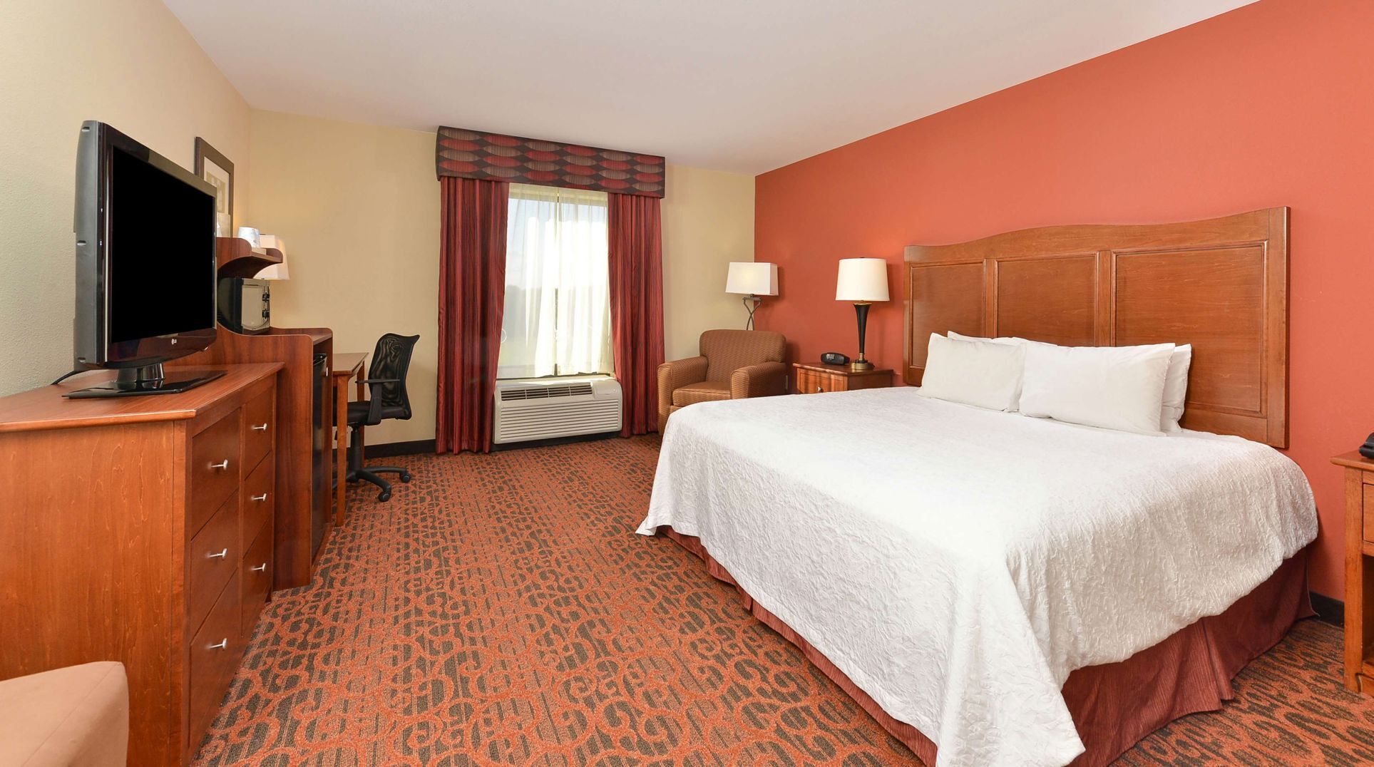 A hotel room with a bed, a desk with a TV, a chair, a lamp, and a window with curtains.