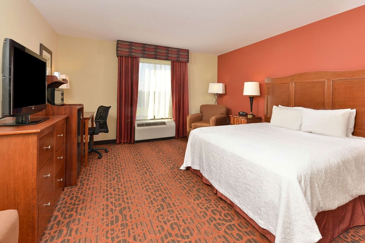 A hotel room with a bed, a desk with a TV, a chair, a lamp, and a window with curtains.