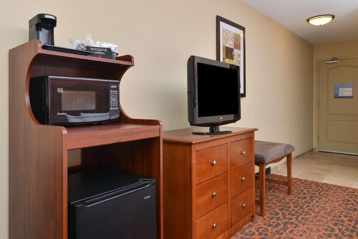 A hotel room with a wooden desk, a microwave, a television, a refrigerator, a bench, and a patterned carpet.