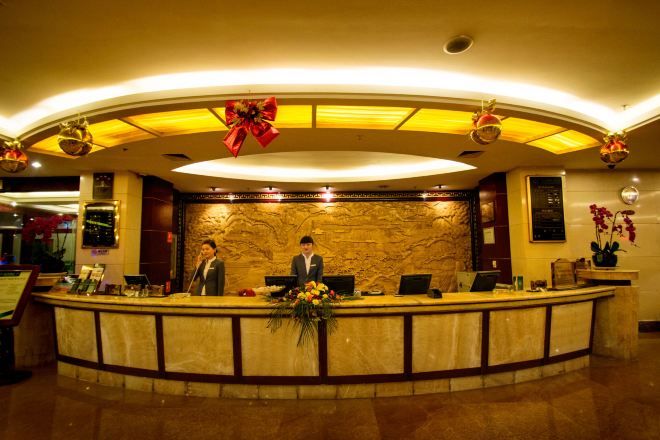 A hotel reception area with a marble counter, two receptionists, a large red bow decoration, and a wall-mounted screen.