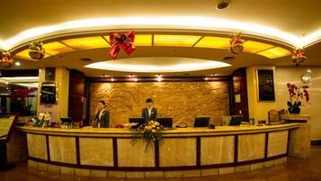 A hotel reception area with a marble counter, two receptionists, a large red bow decoration, and a wall-mounted screen.