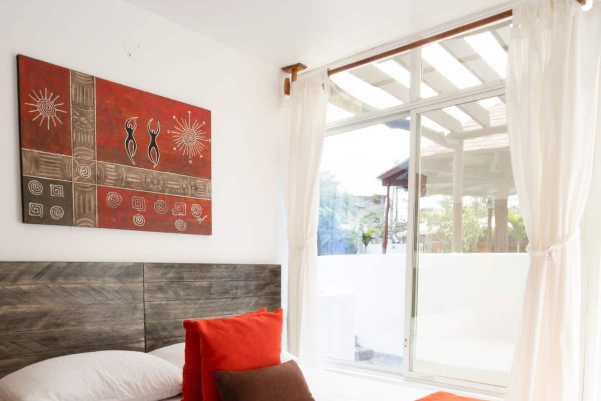 Une chambre d’hôtel avec un grand lit, du linge de lit blanc, un coussin décoratif rouge et marron, une tête de lit en bois, une couverture rouge, une œuvre d’art murale et une grande fenêtre avec des rideaux.