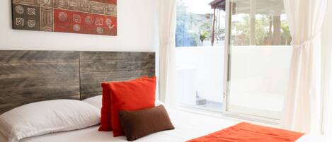 Habitación de hotel con una cama grande, ropa de cama blanca, un cojín decorativo rojo y marrón, un cabecero de madera, una colcha roja, una obra de arte en la pared y un ventanal con cortinas.