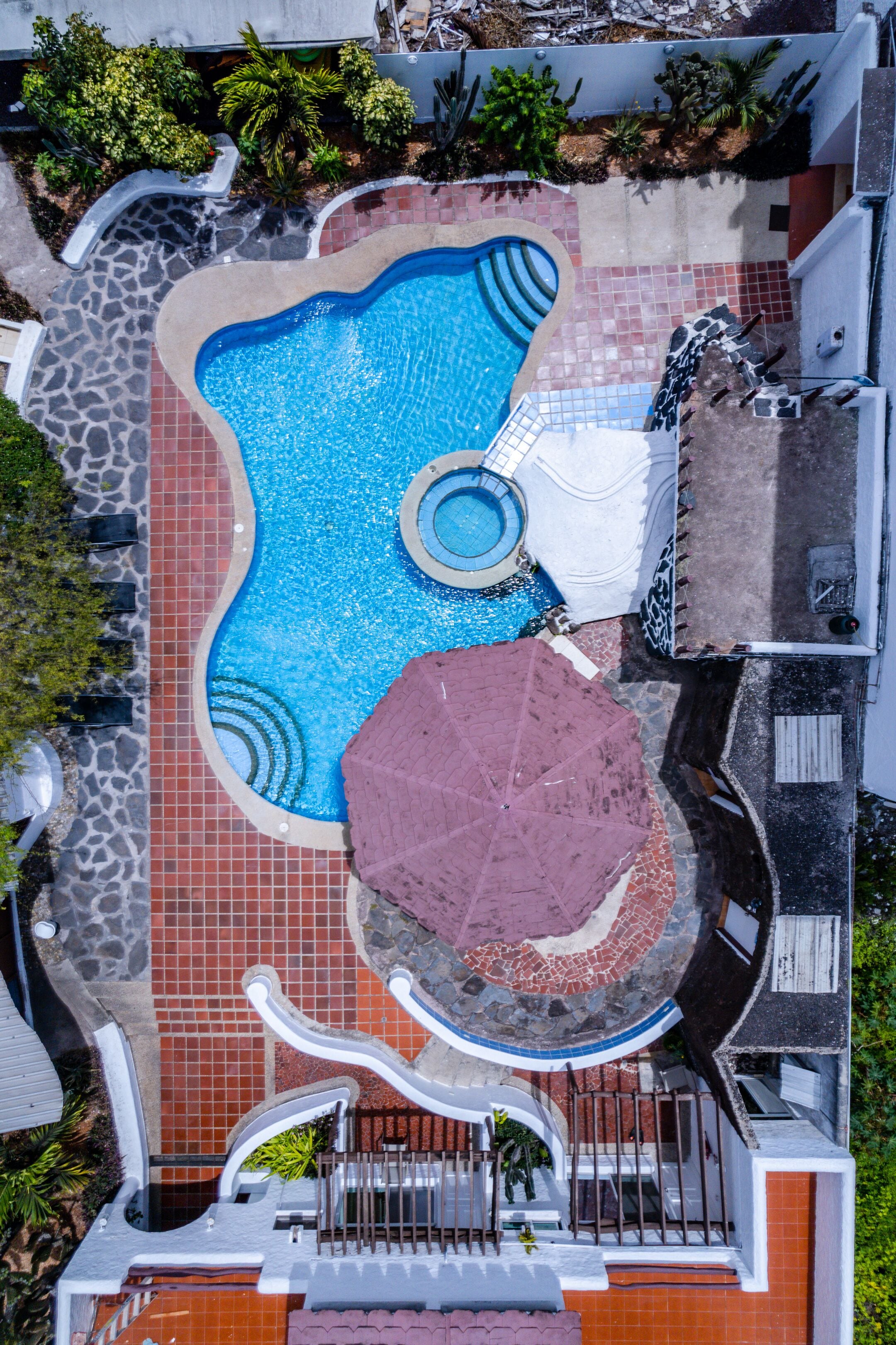 Vista aérea de uma piscina com formato singular, rodeada por um pátio de pedra e tijolo, e adjacente a um edifício com entrada protegida por portão.
