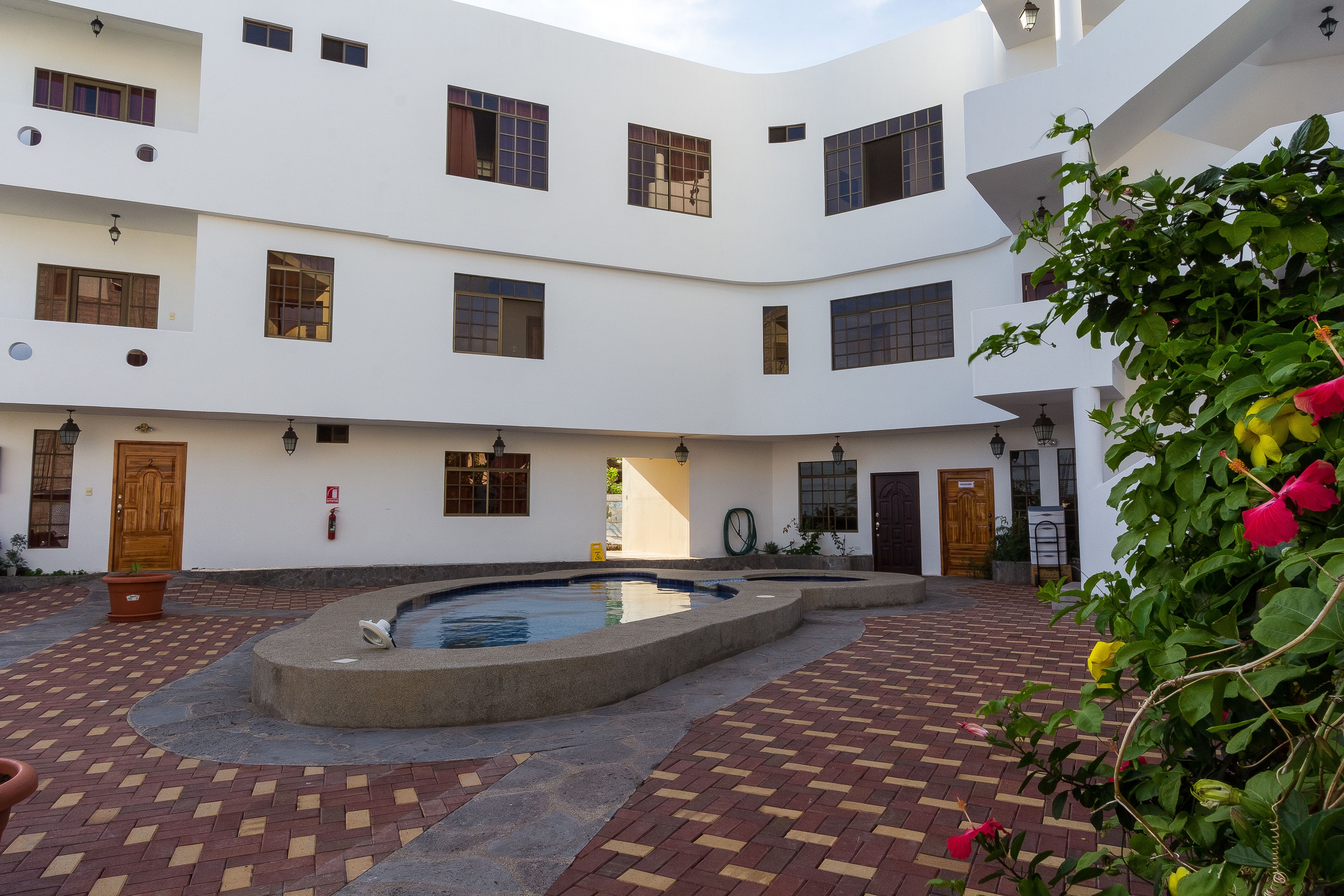 Un patio con piscina, suelo de ladrillo y un edificio blanco con varias ventanas y puertas.