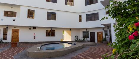 Un patio con piscina, suelo de ladrillo y un edificio blanco con varias ventanas y puertas.