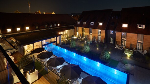 A swimming pool with lounge chairs and umbrellas, surrounded by buildings with multiple windows and a wooden deck. - Hotel Schloss Reinach (Freiburg im Breisgau)