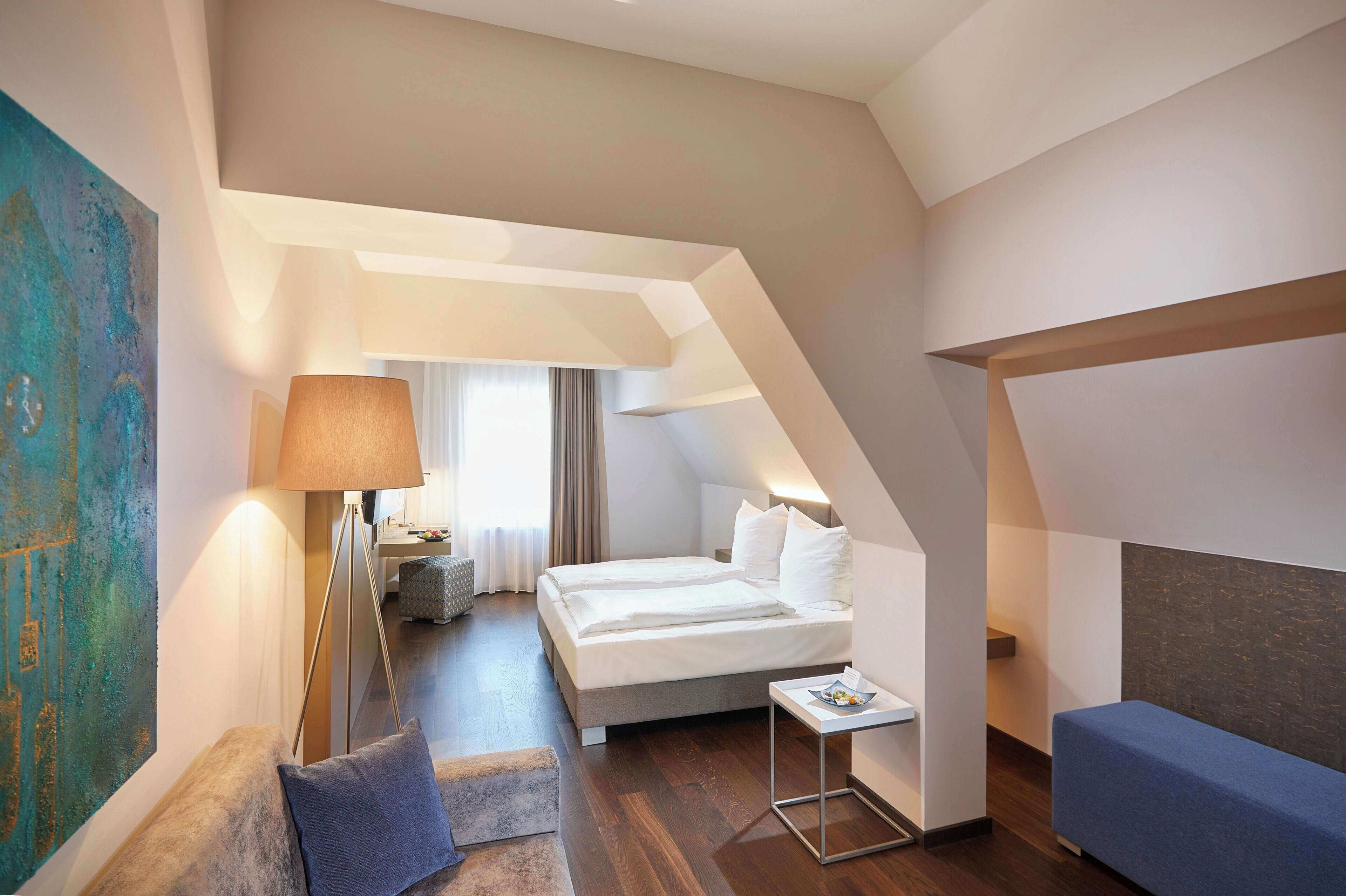 Ett modernt hotellrum med en säng på ett loft, ett sängbord, en lampa och en blå bänk.