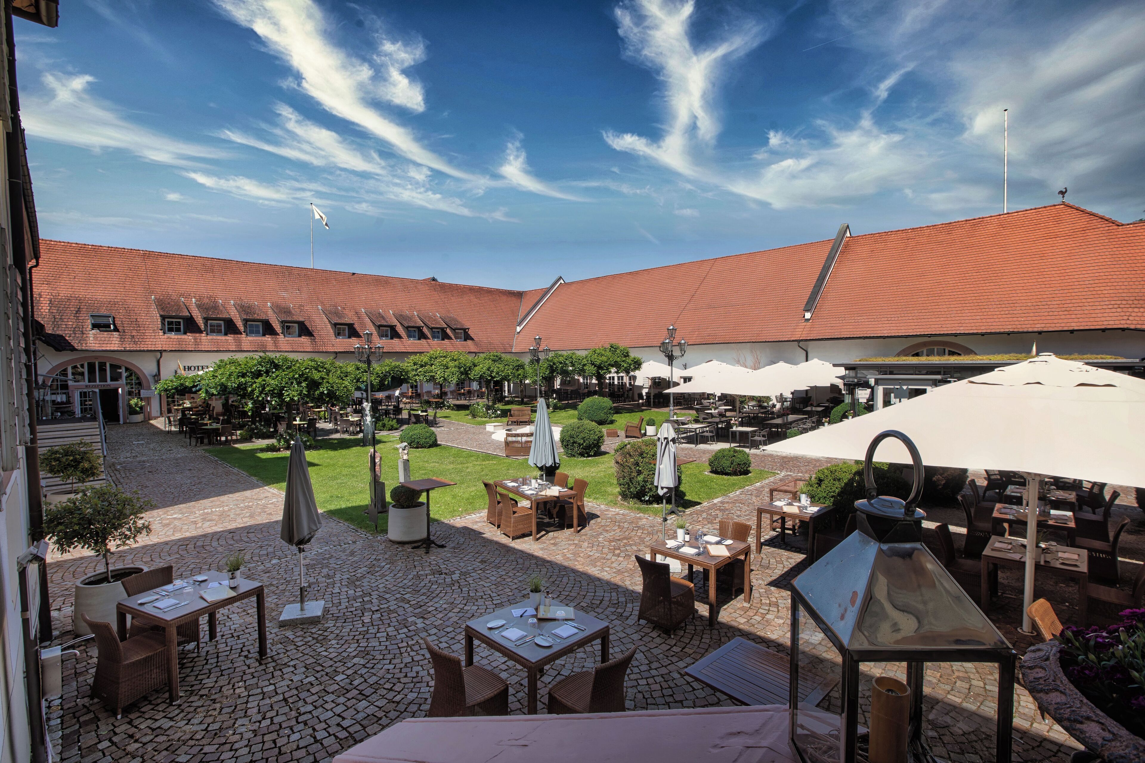 An outdoor patio area with tables and chairs, a garden, and a building with a red roof.