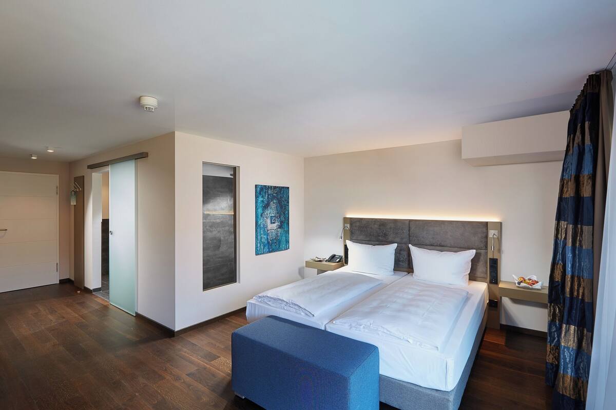 Ein Hotelzimmer mit einem großen Bett, einem Nachttisch mit Lampe, einem Bild an der Wand und Blick auf den Flur.