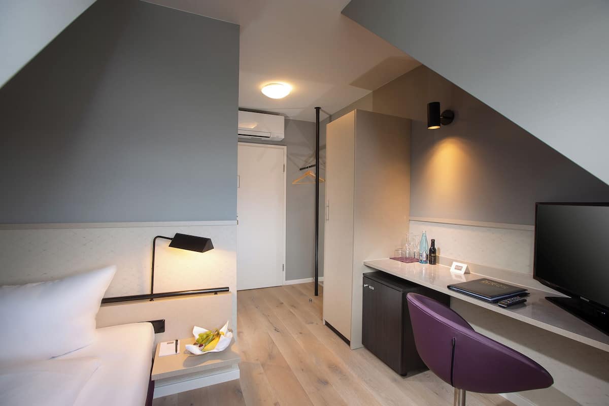 Ein kompaktes Hotelzimmer mit Bett, Nachttisch, Lampe und einer kleinen Küchenzeile mit Fernseher, Kühlschrank und Barhocker.