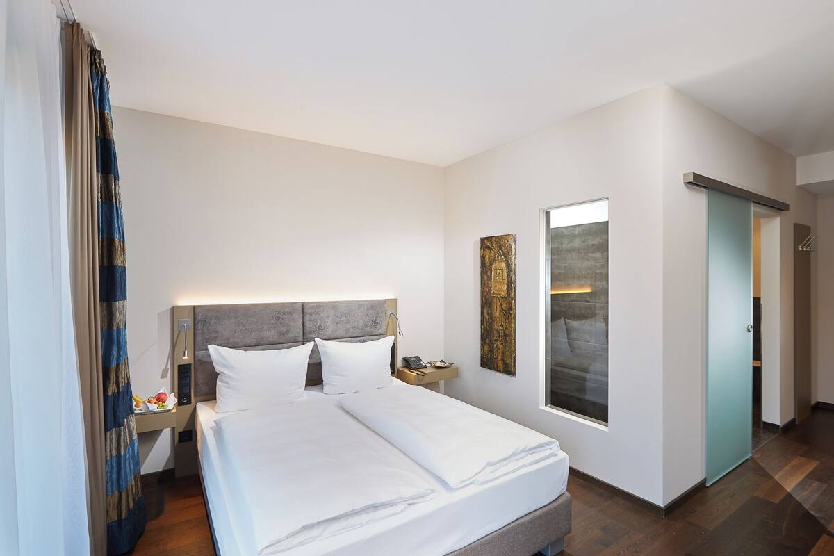 Ein Hotelzimmer mit Bett, Nachttisch, Lampe, Kunstwerk und Blick nach draußen.