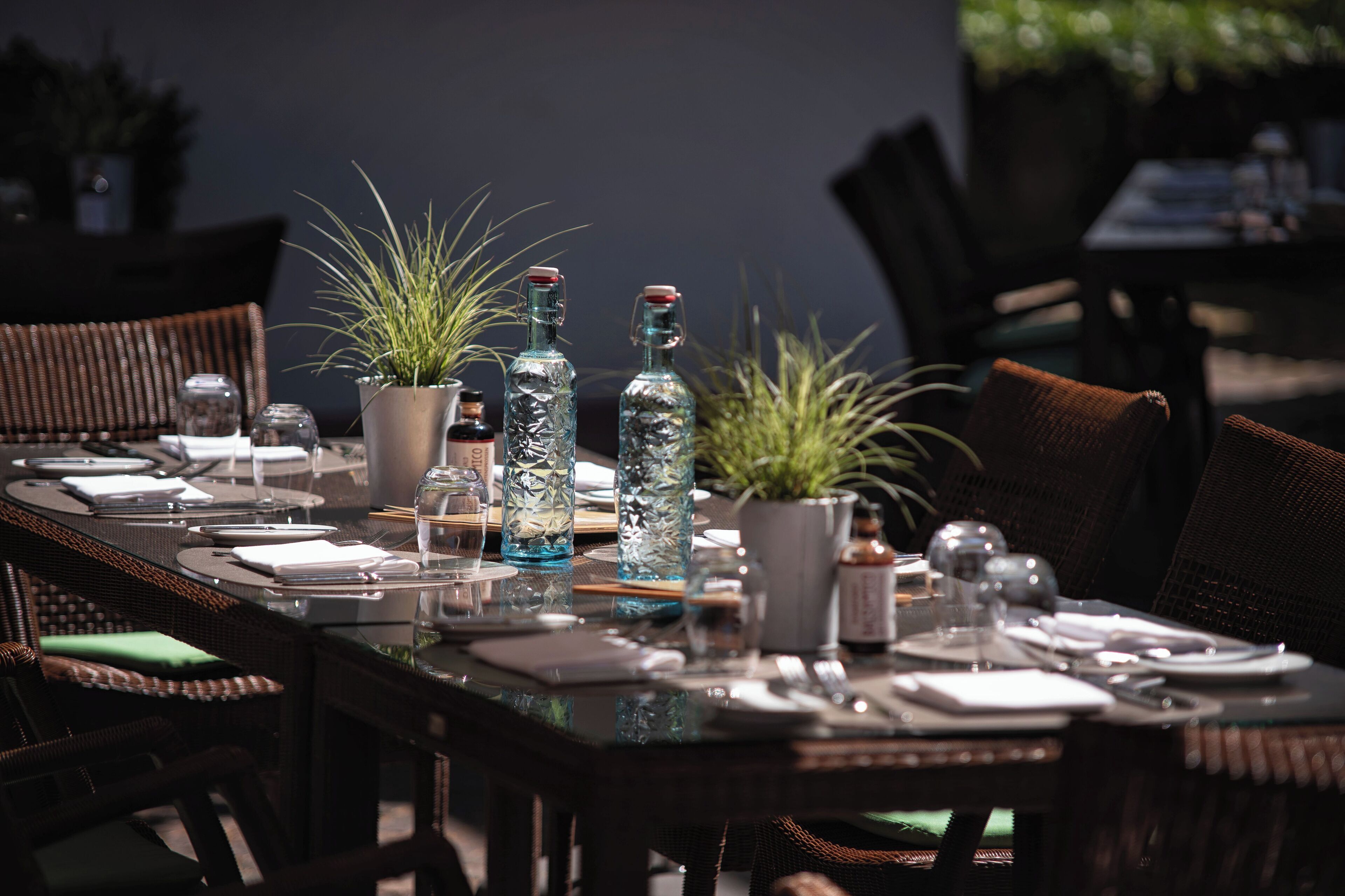 Una mesa de comedor al aire libre con sillas de mimbre, plantas en macetas y botellas de vidrio transparente.