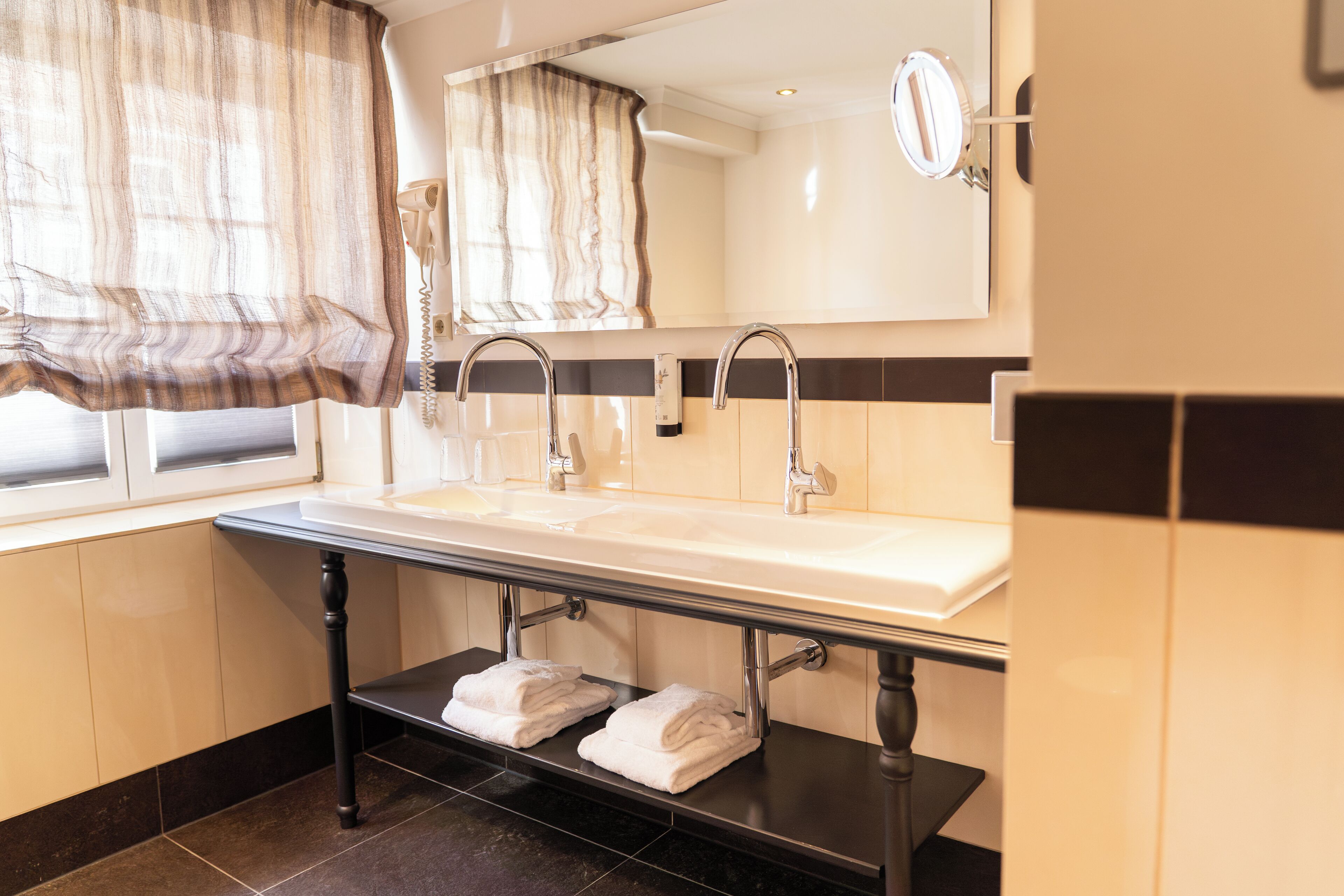 Une salle de bain avec un double lavabo, un miroir, un porte-serviettes et une fenêtre avec des rideaux.