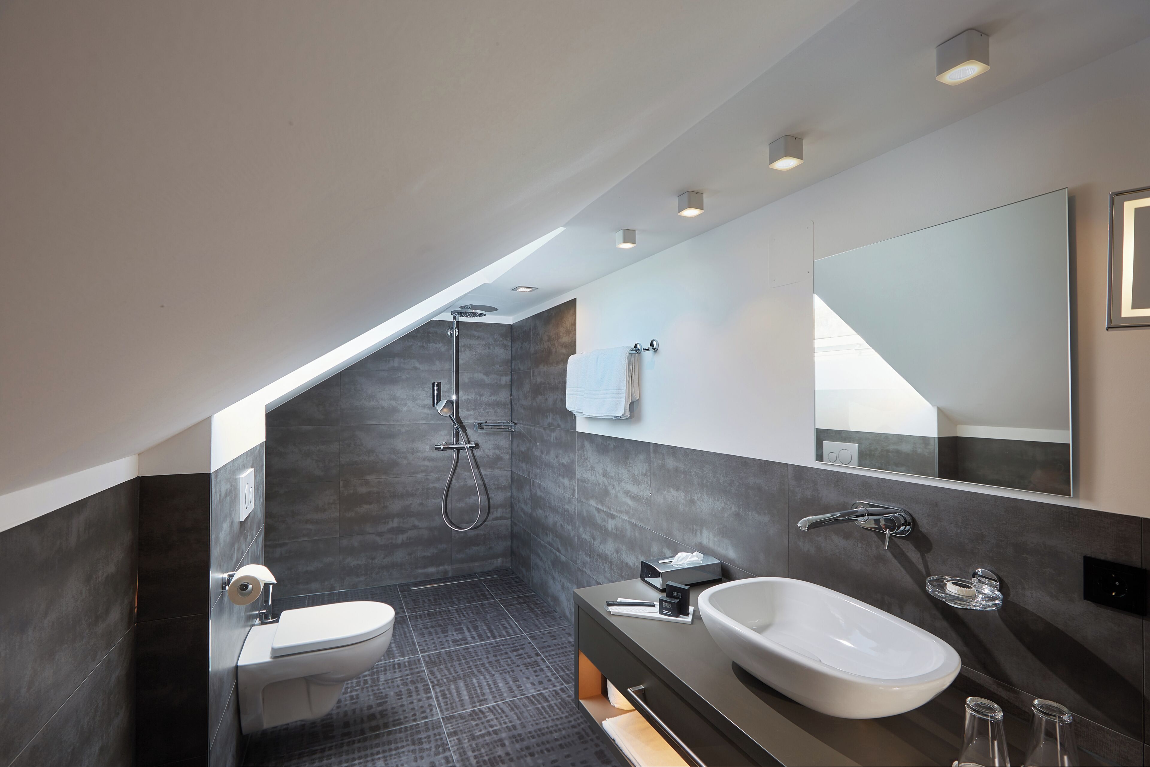 Un baño moderno con ducha, inodoro y lavamanos, con baldosas oscuras e iluminación empotrada.