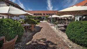 A cobblestone pathway leading to outdoor seating areas with umbrellas, surrounded by greenery and a building with a tiled roof. - Hotel Schloss Reinach (Freiburg im Breisgau)