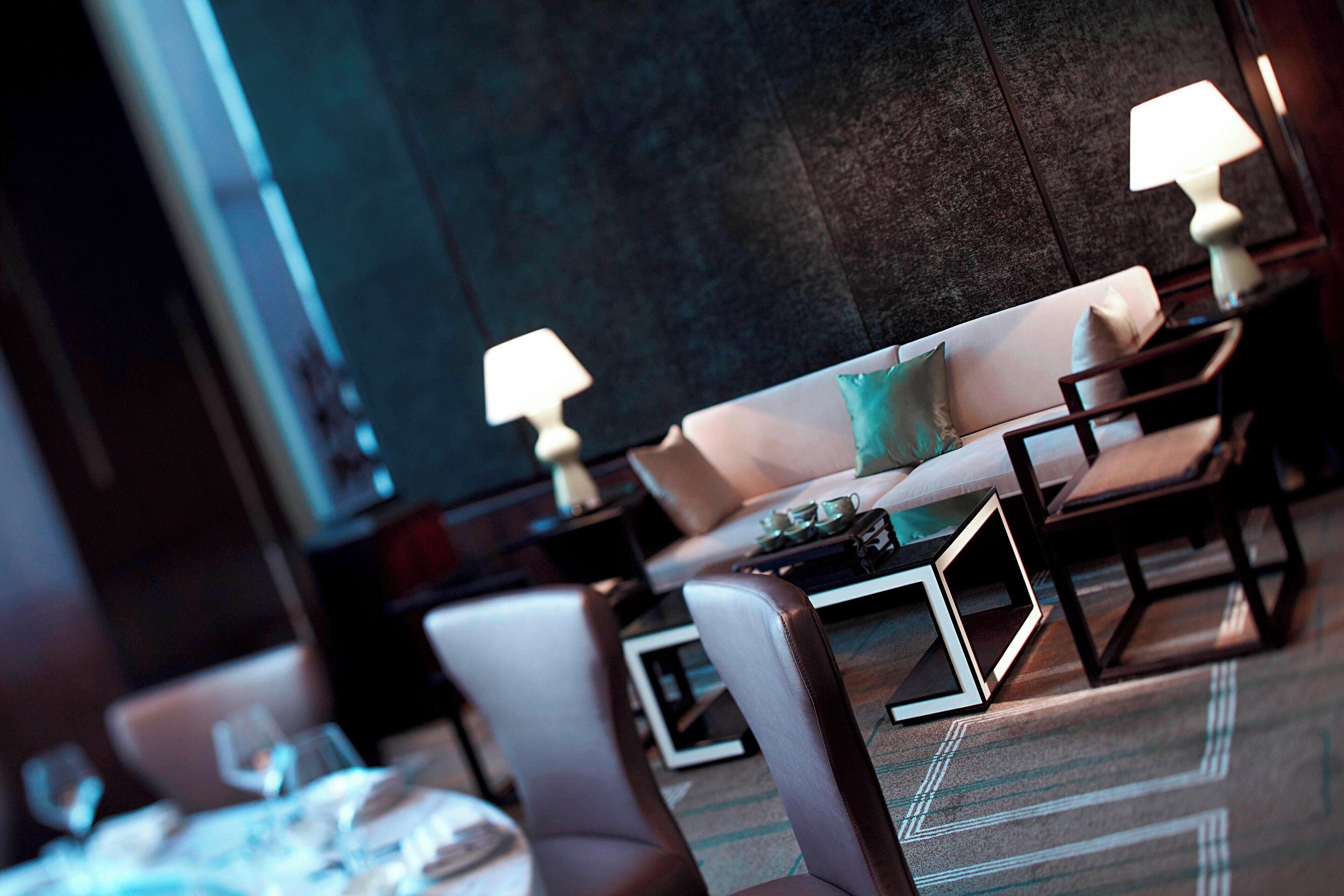 A modern hotel room with a sofa set, a dining table with chairs, and a lamp on a side table.
