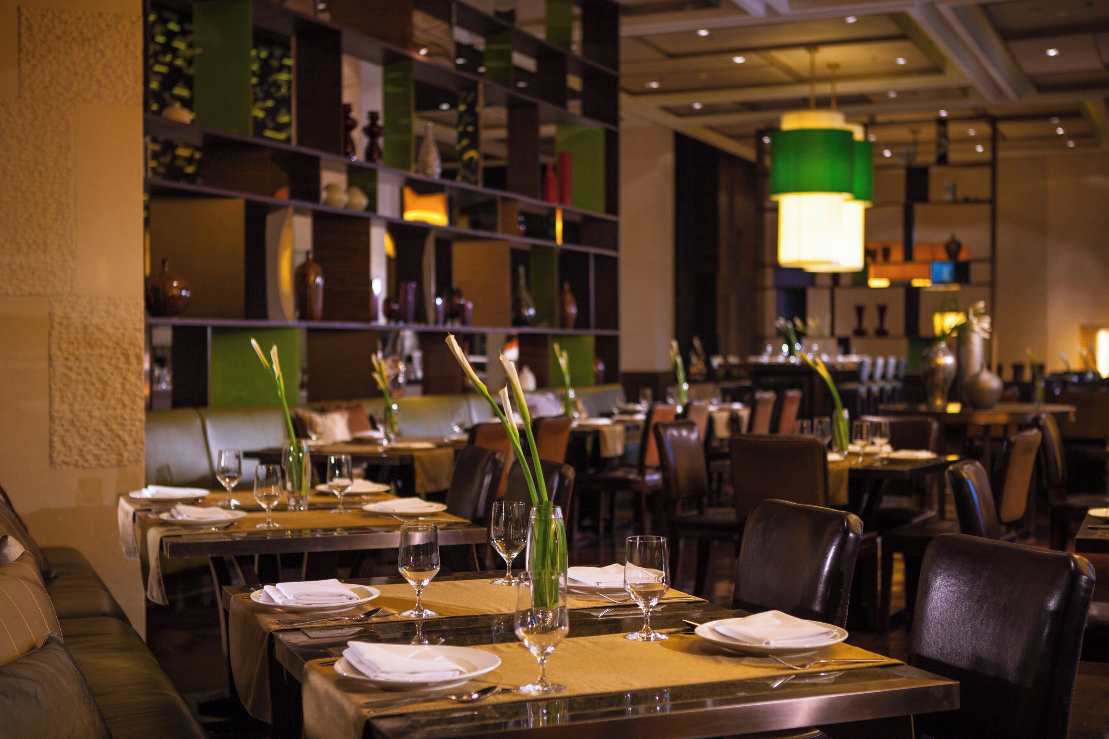 A restaurant interior with tables set for dining, featuring green decorative vases and a warm ambient lighting.