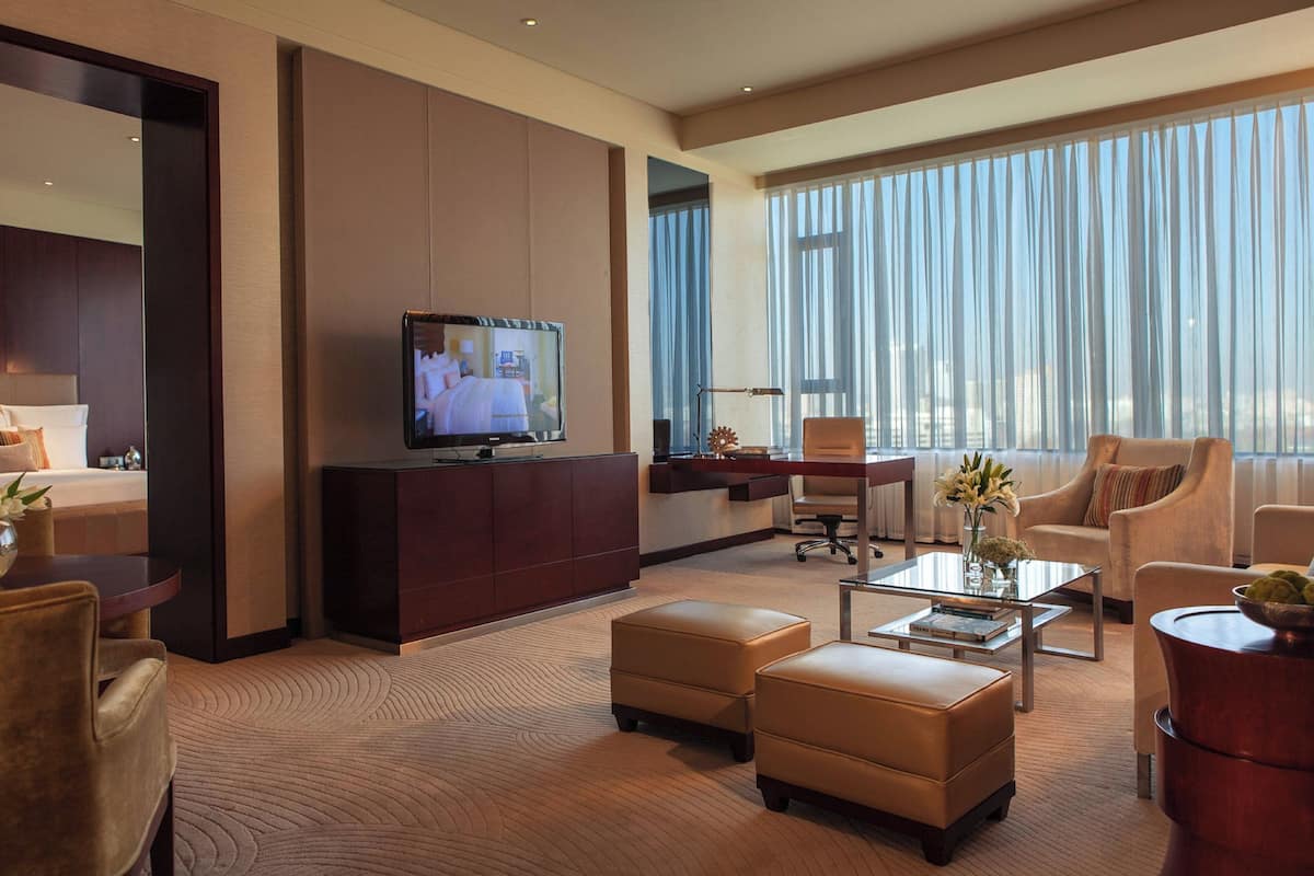 A modern hotel room with a large TV, comfortable seating, a desk, and a view of the city through large windows.