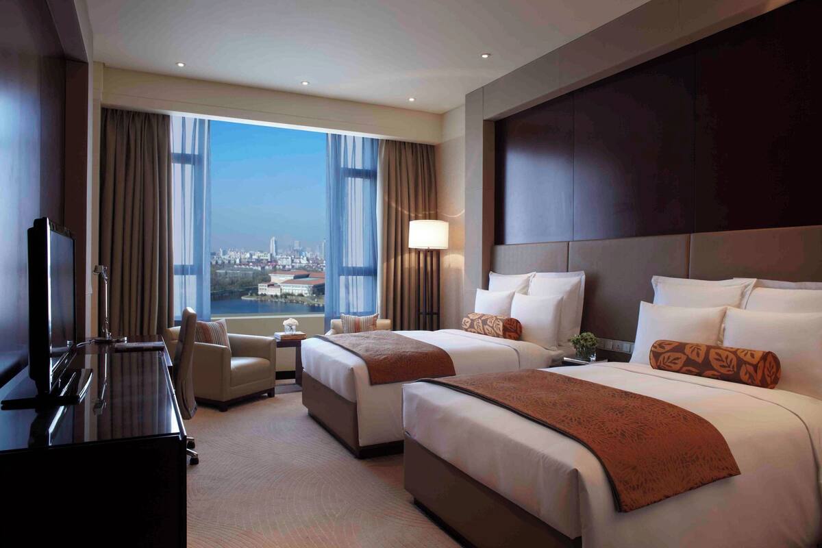 A hotel room with two beds, a desk, a chair, and a large window offering a city view.