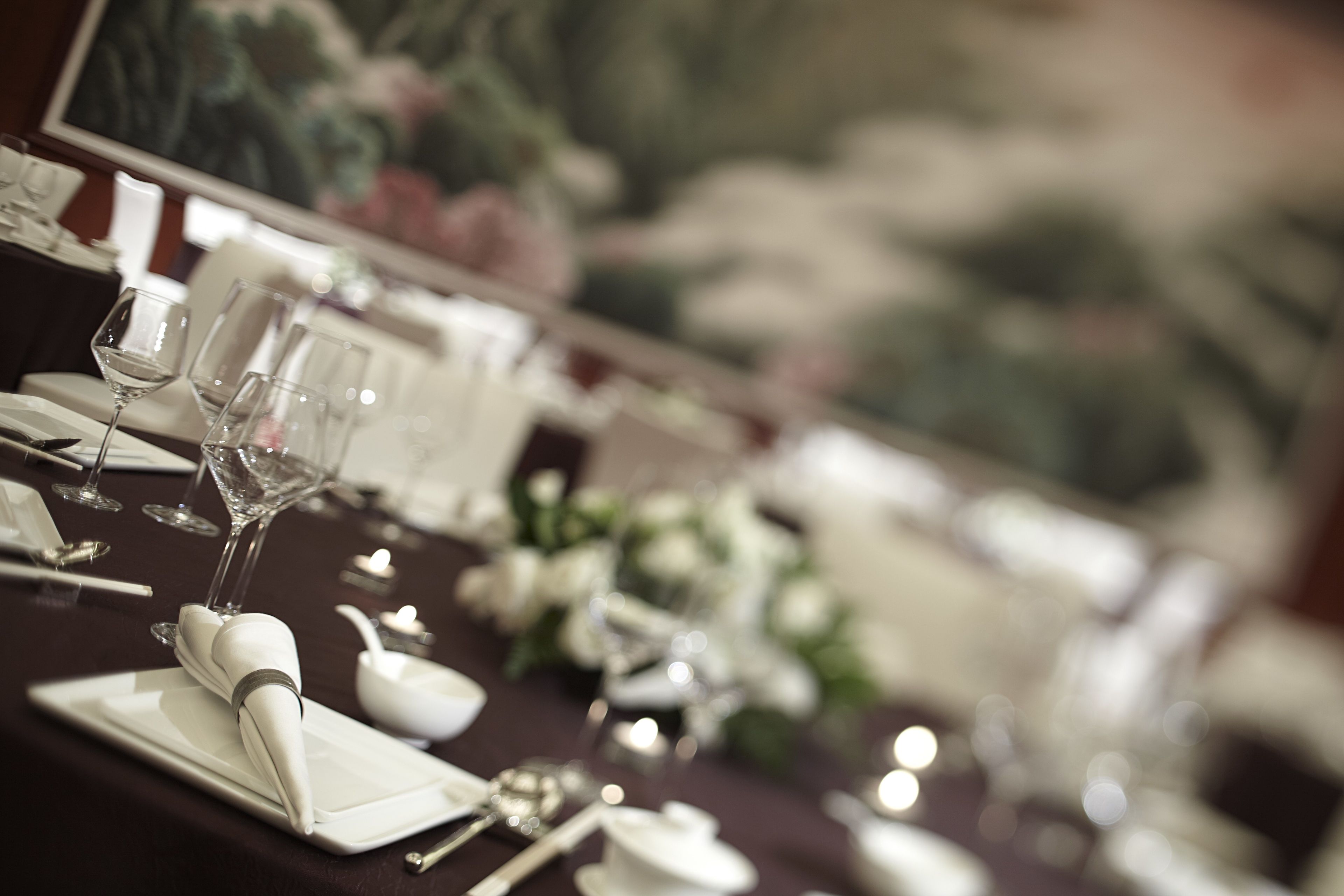 A dining table set with white tableware and glassware, with a painting in the background.