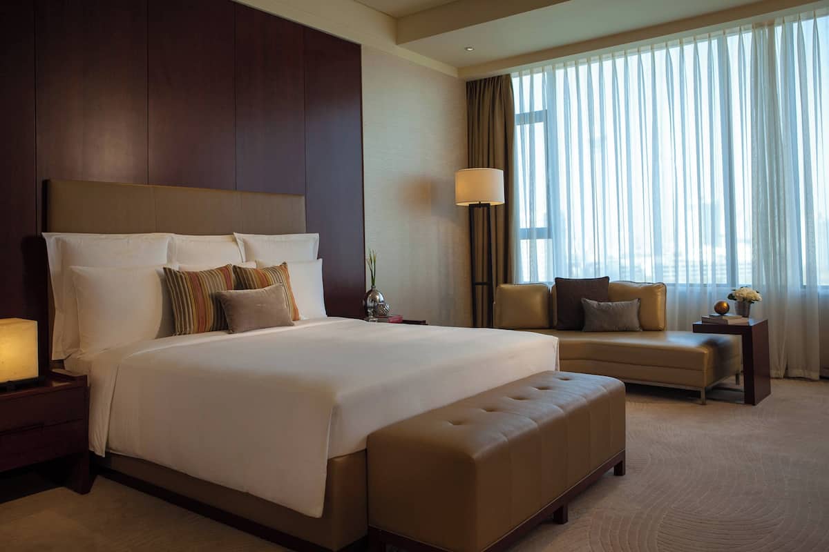 A large bed with multiple pillows, a bedside table with a lamp, a sofa, and a window with curtains.