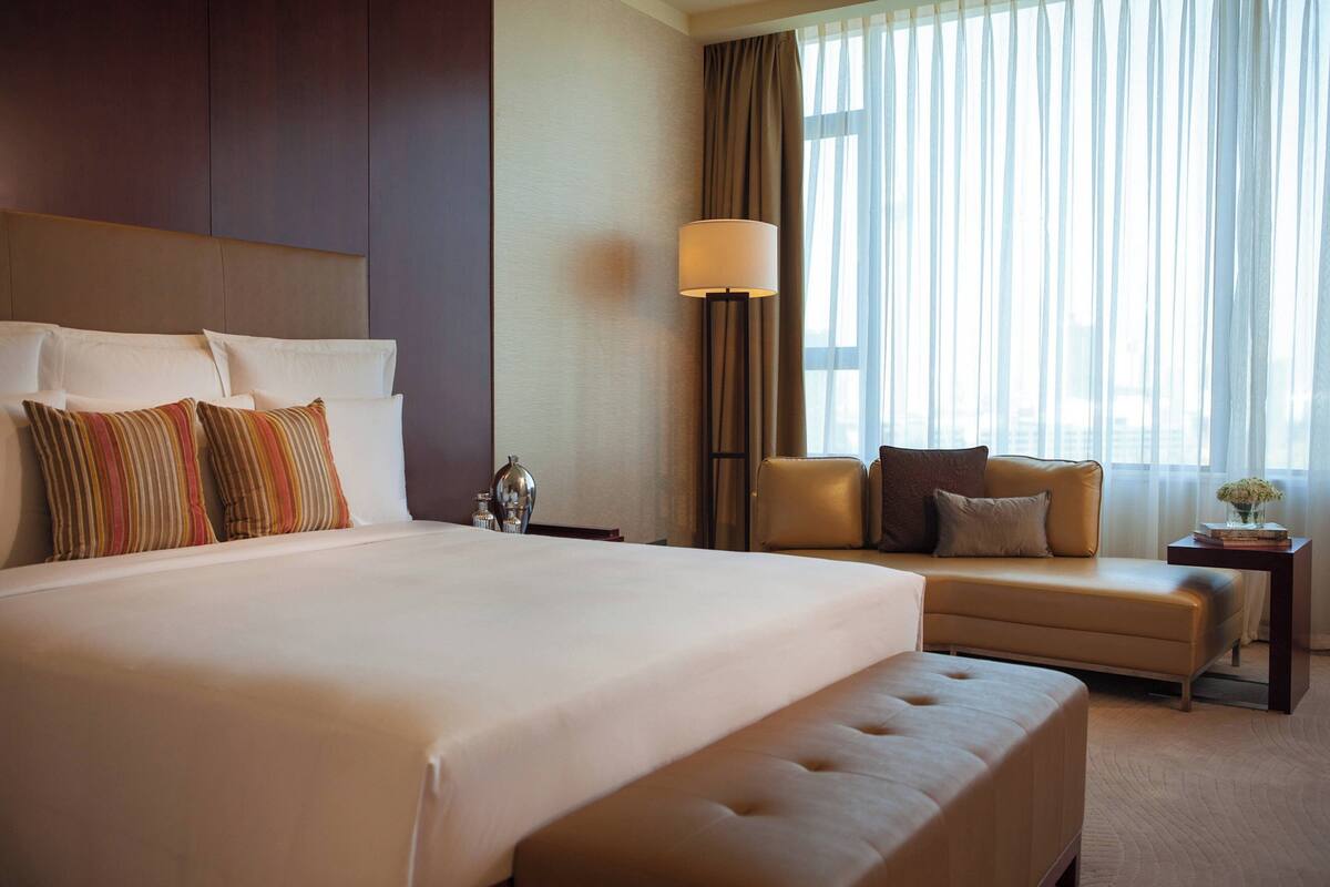 A hotel room with a large bed, a sofa, a lamp, and a view of the city through the window.