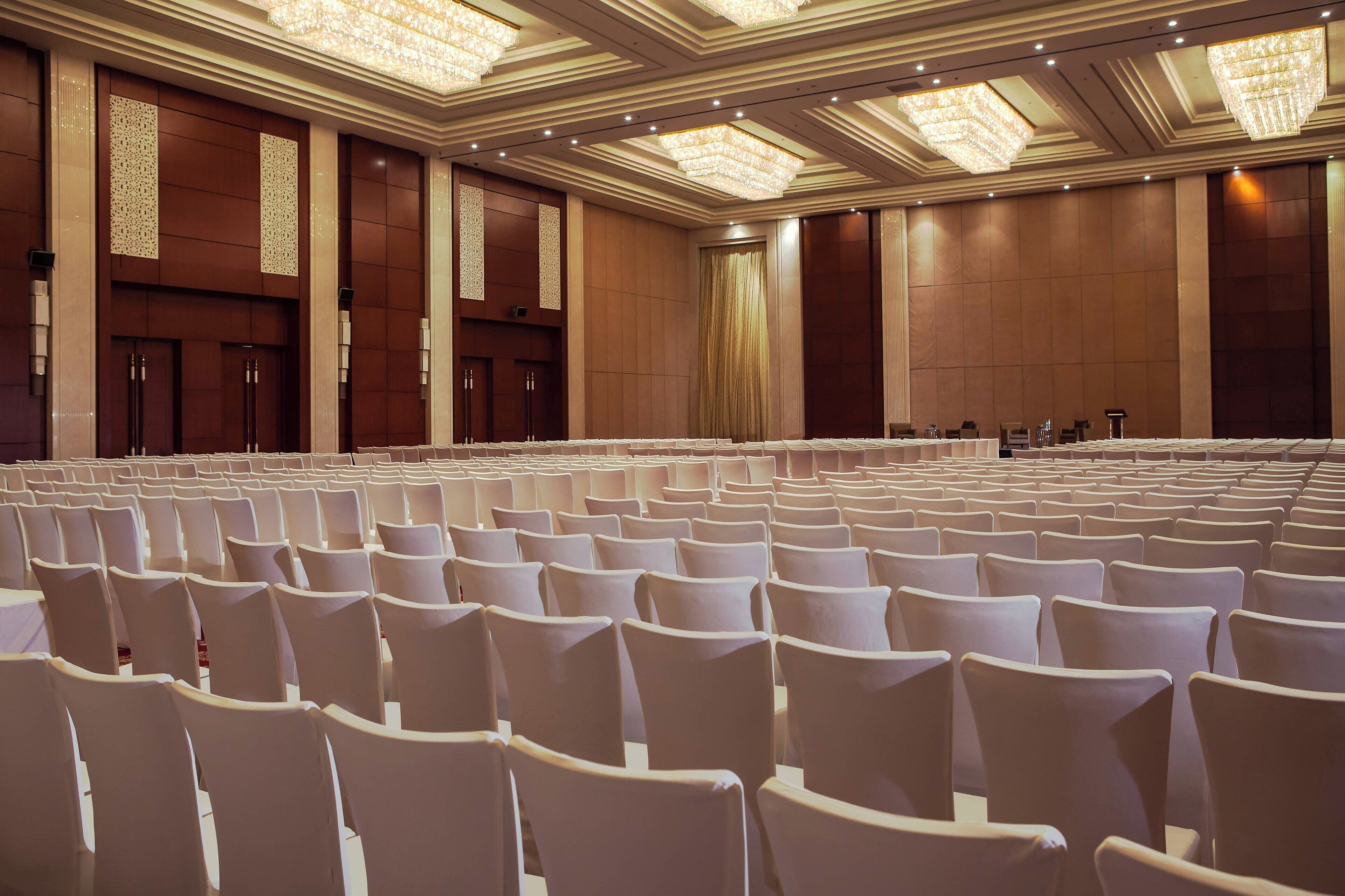 Un salón de conferencias con hileras de sillas blancas, un escenario con un atril y candelabros en el techo.