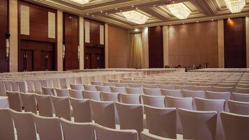 Un salón de conferencias con hileras de sillas blancas, un escenario con un atril y candelabros en el techo.