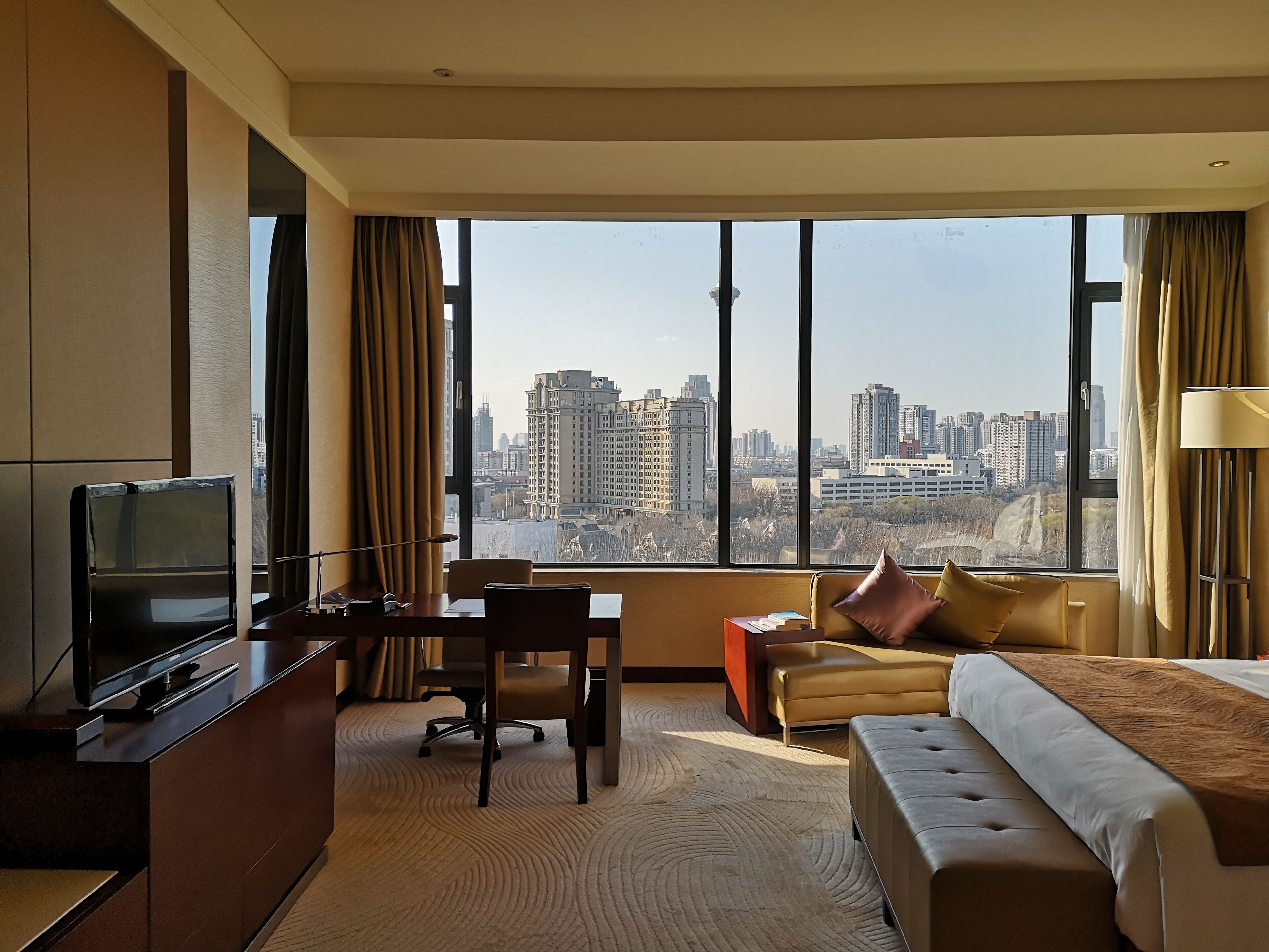 A hotel room with a large window, a bed, a desk, and a television.