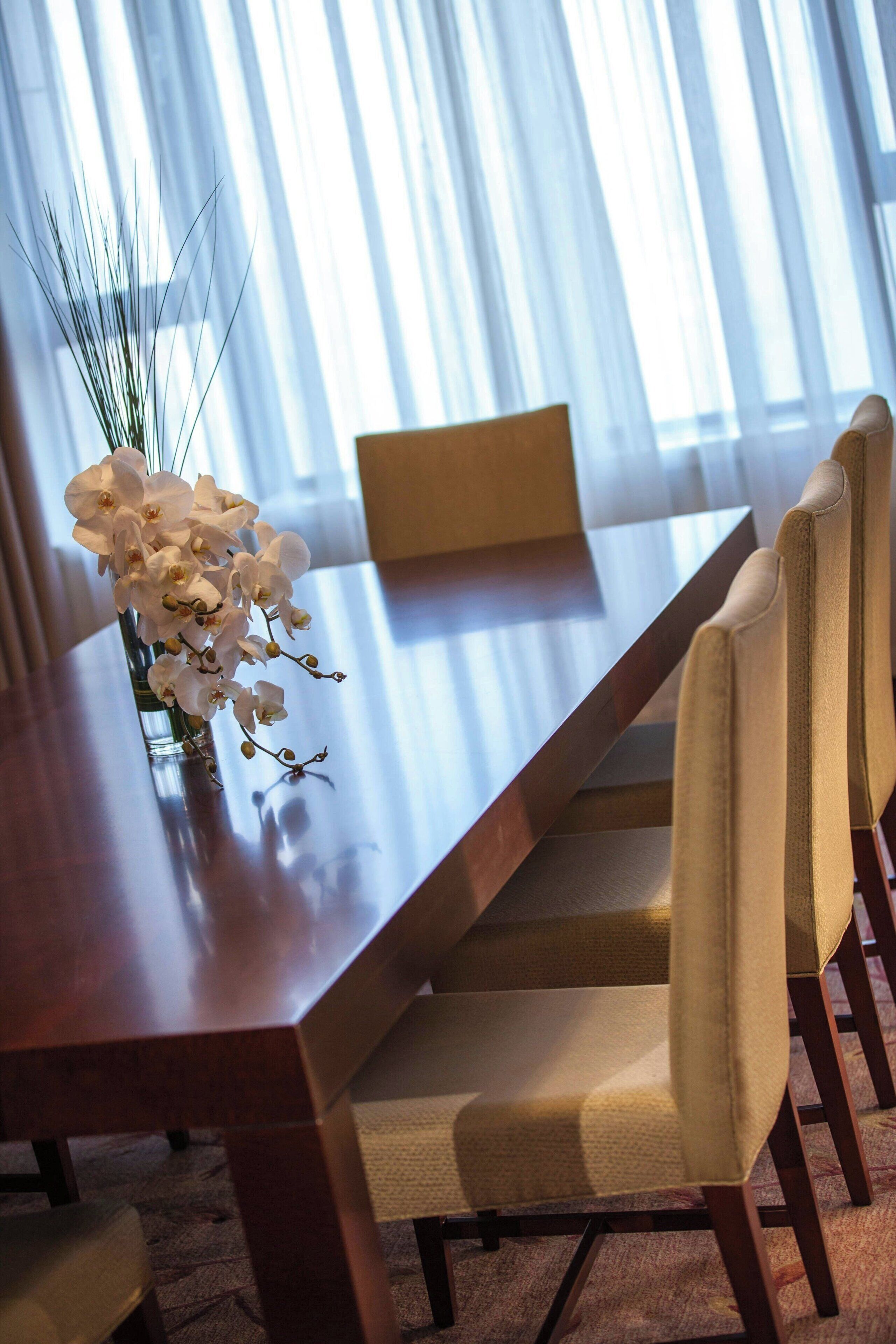 A dining table with chairs, a vase with white flowers, and a window with sheer curtains.