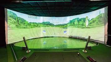 A golf simulation setup with a large screen displaying a virtual golf course, a green practice mat, and electronic score tracking devices.