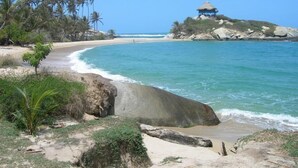 A beach with clear turquoise waters, a thatched-roof structure on a rocky outcrop, and palm trees. - Hotel Bahia Blanca (Santa Marta)