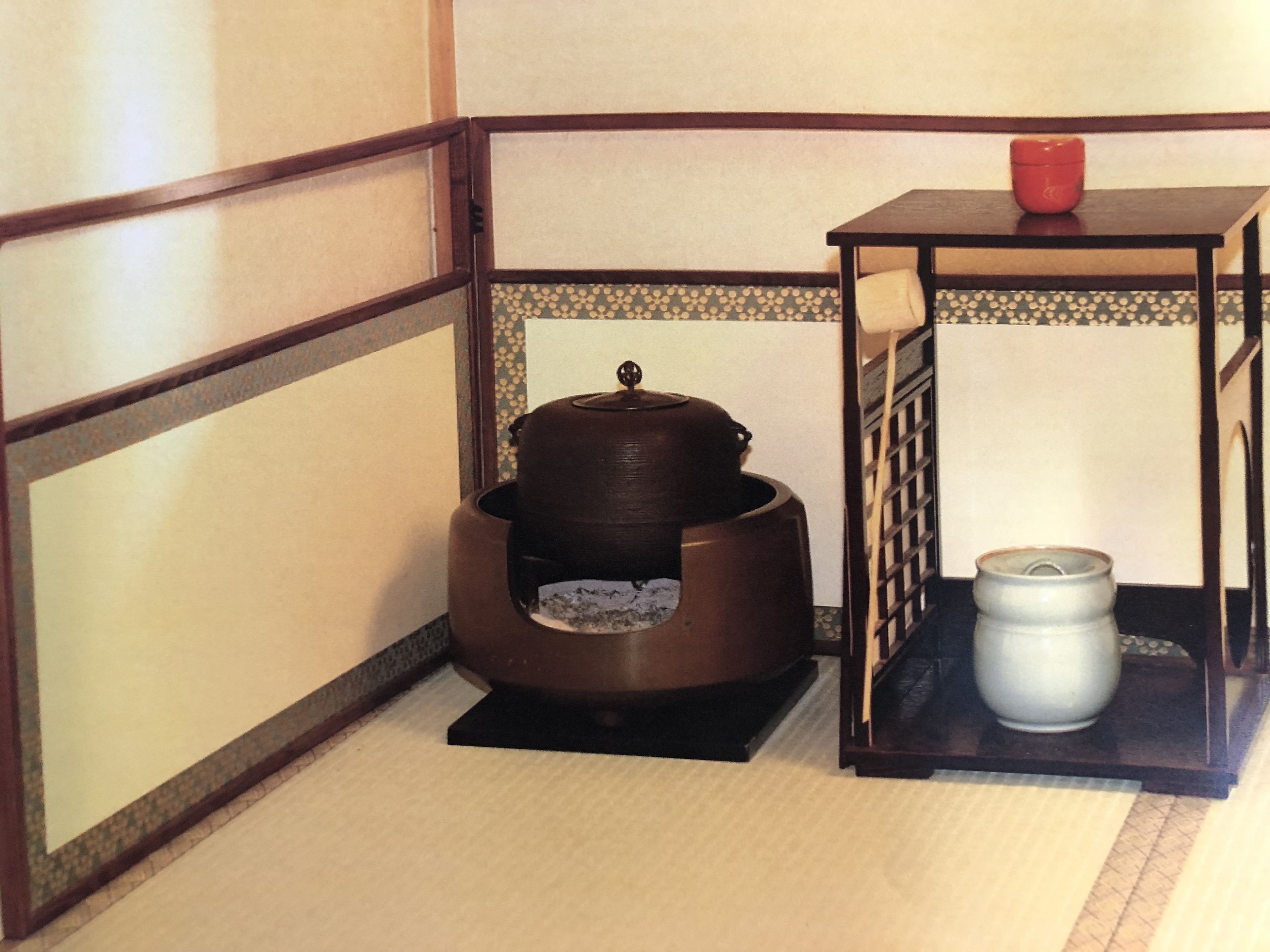 A traditional Japanese room with tatami flooring, a low table with a red container, a large ceramic pot, and a white container on a small table.