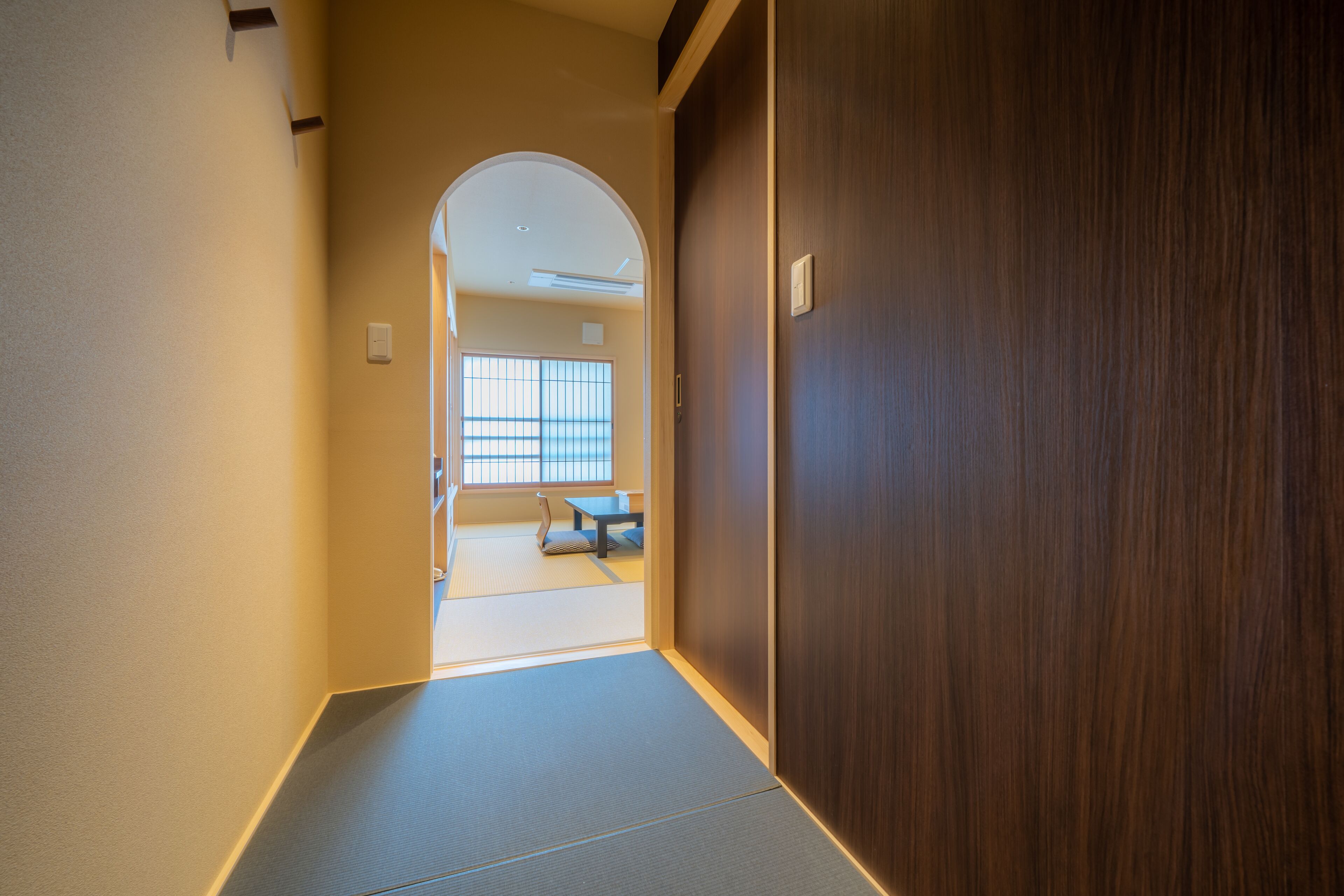 Un couloir avec une porte en bois, un sol recouvert de tatamis et une pièce de style japonais traditionnel visible à travers une porte en arc de cercle.