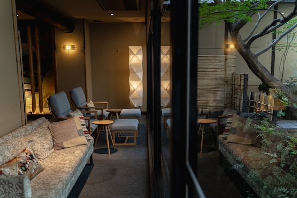 A cozy indoor seating area with sofas, chairs, and tables, featuring a decorative wall and a tree outside.