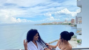 Dos personas en un balcón con vista al mar y al paisaje urbano.