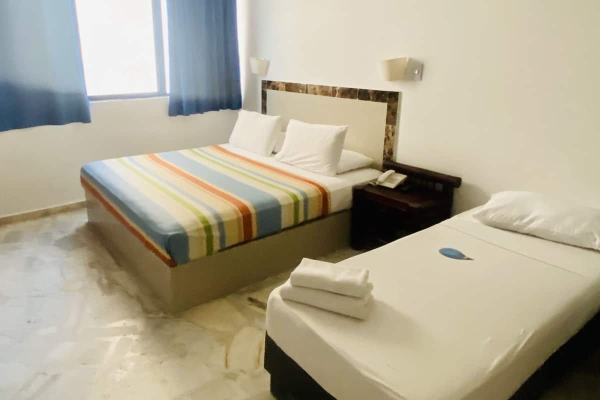 A hotel room with two beds, one with a striped bedspread and the other with a plain white bedspread. There is a nightstand with a phone and a lamp between the beds.
