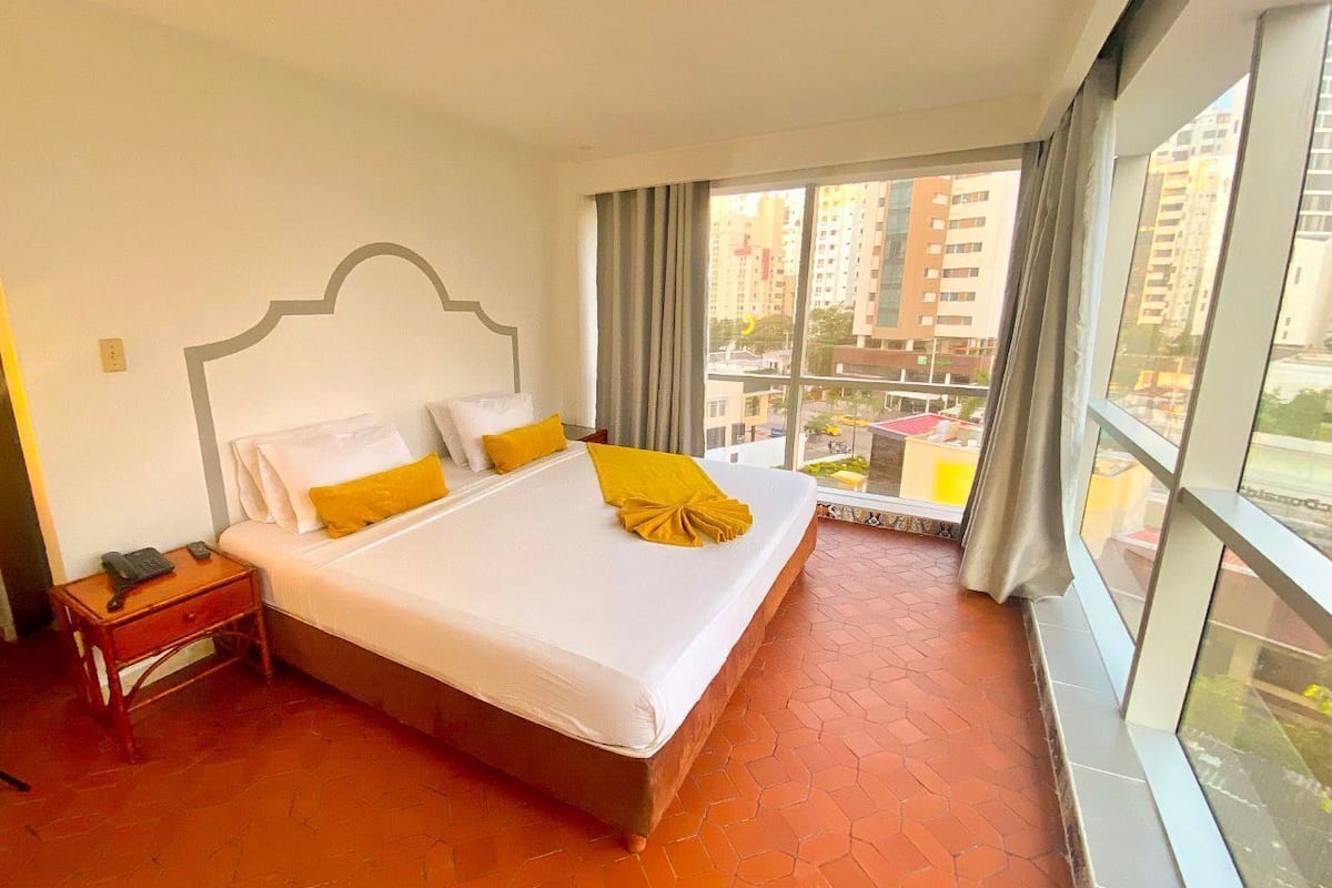 A hotel room with a large bed, a nightstand, and a view of the city through large windows.